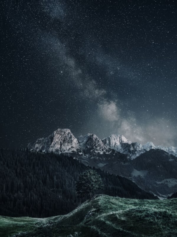 A breathtaking night scene with a star-filled sky and the Milky Way above rugged, snow-capped mountains. Below, a lush green valley with a solitary tree enhances the landscape's serene beauty.