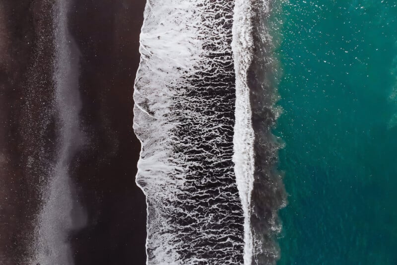 Aerial view of a coastline where dark sand meets turquoise ocean waves. The white foam of the waves creates a contrasting border between the land and sea, showcasing the striking difference in colors and textures.