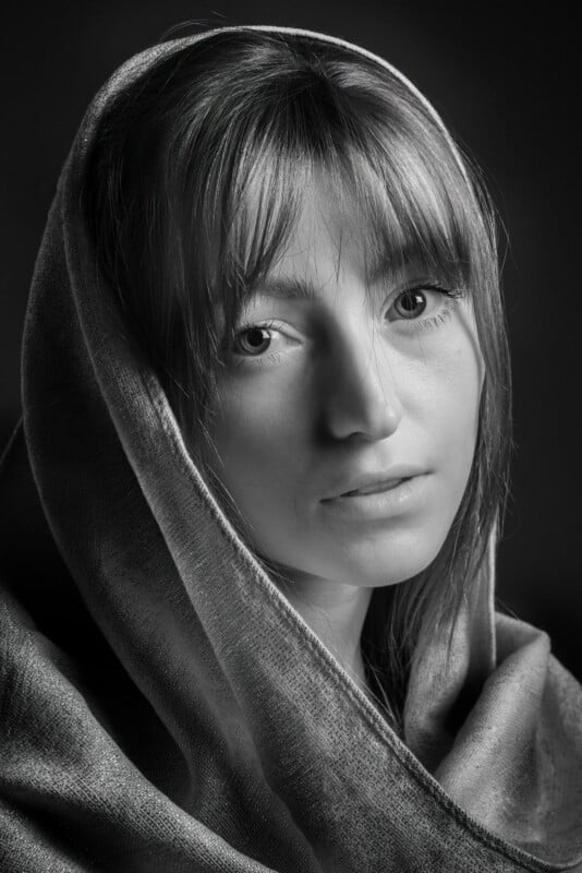 Monochrome portrait of a woman exuding a soft demeanor, partially veiled by a textured shawl draped over her head. Her gaze meets the camera directly, while soft illumination emphasizes her features against a dark backdrop.