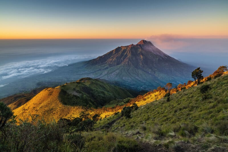 An awe-inspiring vista of a mountain at dusk, featuring a path winding through emerald hills and valleys. The sky displays a gradient, shifting from deep orange to blue, and clouds embrace the mountainside, enhancing the tranquil landscape.