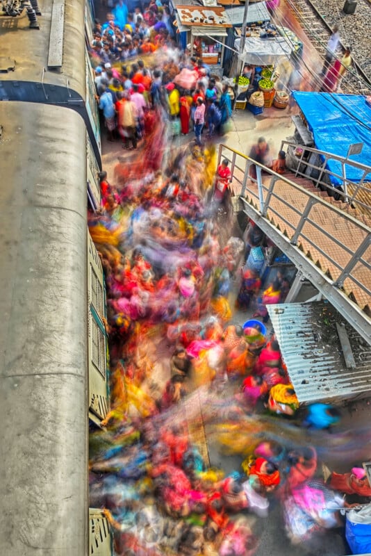 Blurry depiction of a bustling railway station platform with a train on the left. A vibrant assembly of individuals is in motion, forming a lively swirl of colors. Market stalls are arranged on the side, with a footbridge visible overhead.