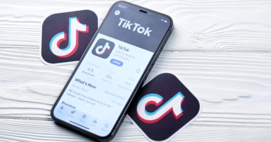 A smartphone displaying the TikTok app page on the screen rests on a wooden surface. Two TikTok logo stickers are placed beside the phone. The app page shows a 4.8-star rating and the "Open" button.