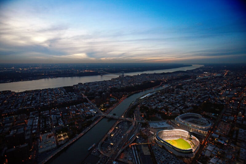 Aerial view of a brightly lit stadium in a cityscape at twilight. The surrounding buildings and roads are illuminated, with a river visible, stretching towards the horizon under a partially cloudy sky.