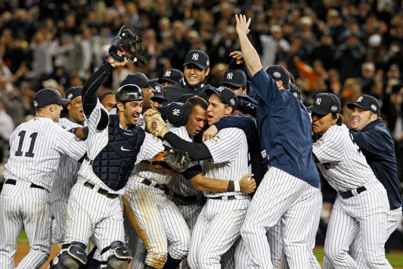 A jubilant baseball team, wearing striped uniforms, celebrates on the field. Players are huddled together, smiling and raising their arms in excitement. The background shows a cheering crowd under stadium lights.