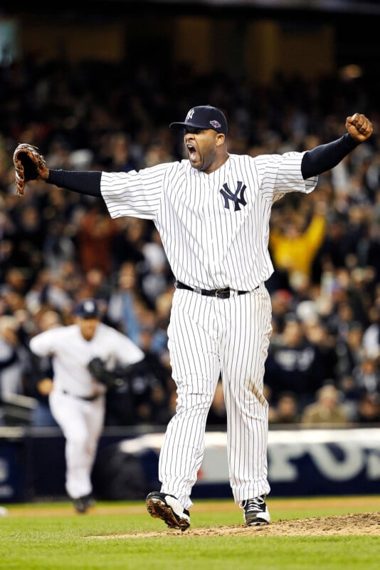 Baseball player in a pinstripe uniform celebrates energetically on the field with arms raised. The crowd is visible in the background, and a teammate runs toward him.