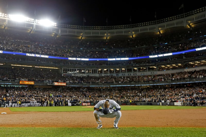 A baseball player crouches on the field at night, surrounded by a packed stadium. Bright lights illuminate the scene, with a large crowd visible in the stands and a "THANK YOU" message displayed above.