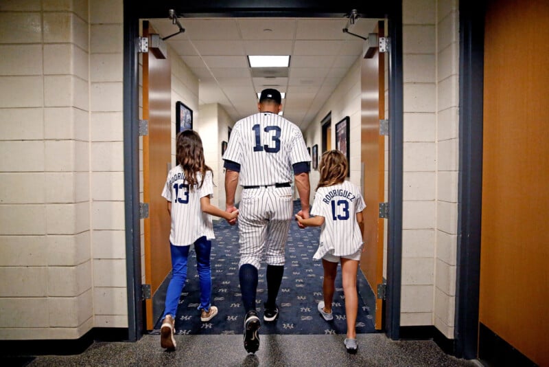 A baseball player in a pinstripe uniform with the number 13 walks down a hallway holding hands with two children, also wearing jerseys with the number 13. The scene captures a family moment in a sports setting.