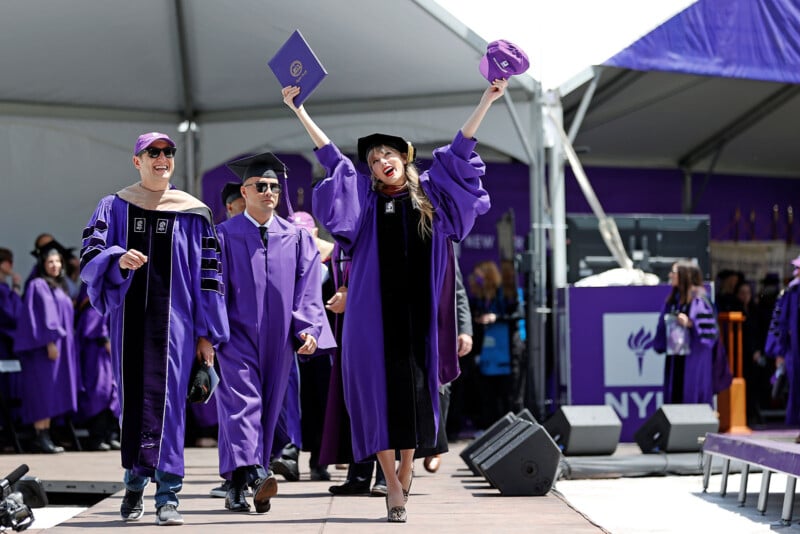 Graduates in purple robes celebrate at a commencement ceremony. One person raises a diploma and cap joyfully, while others smile. They are walking on a stage under a tent with people and banners in the background.