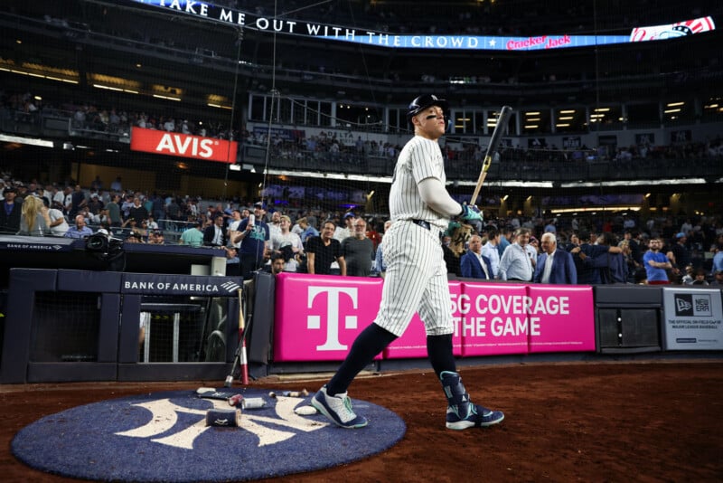 A baseball player in a pinstripe uniform and helmet stands on the field holding a bat. A crowd is visible in the stadium behind him. Vibrant signage from sponsors adorns the background.