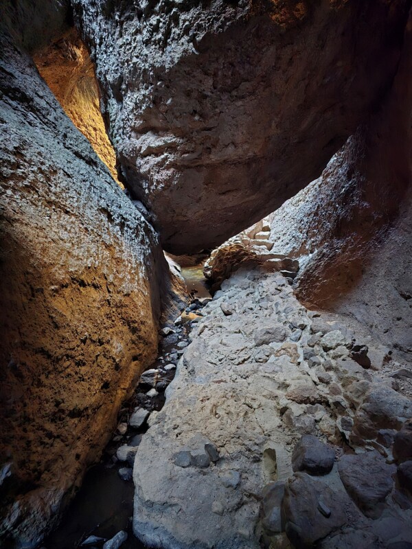 View of a narrow rocky canyon with light illuminating the textured walls. A small stream runs between the rocks, leading to a large boulder overhead. The scene features a mix of rugged rock surfaces and scattered stones.