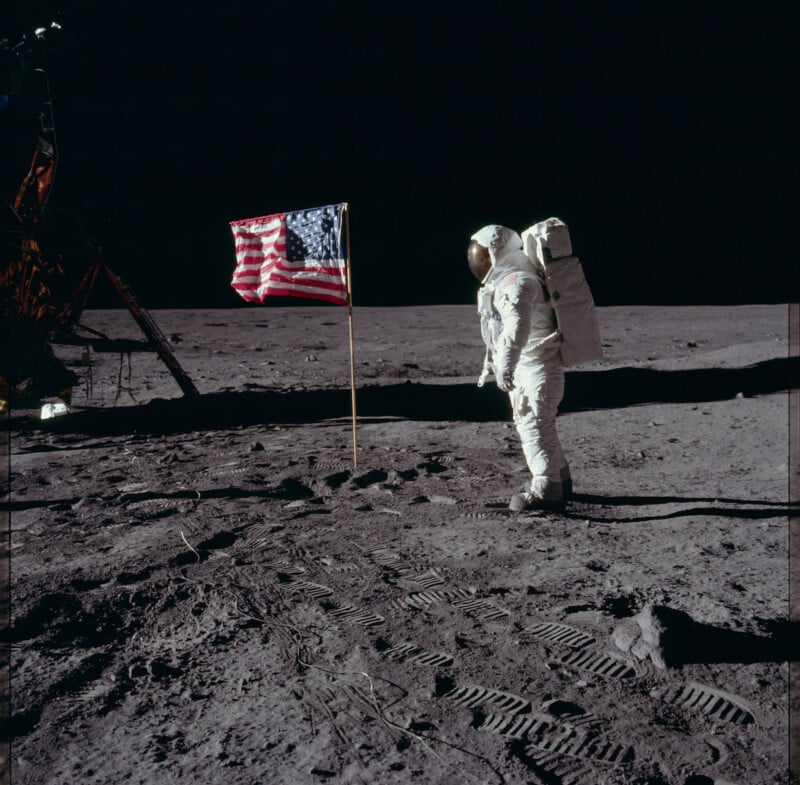 An astronaut stands on the moon's surface, wearing a spacesuit. An American flag is planted nearby, and footprints are visible in the lunar dust. The lunar module is partially visible in the background against the dark space.