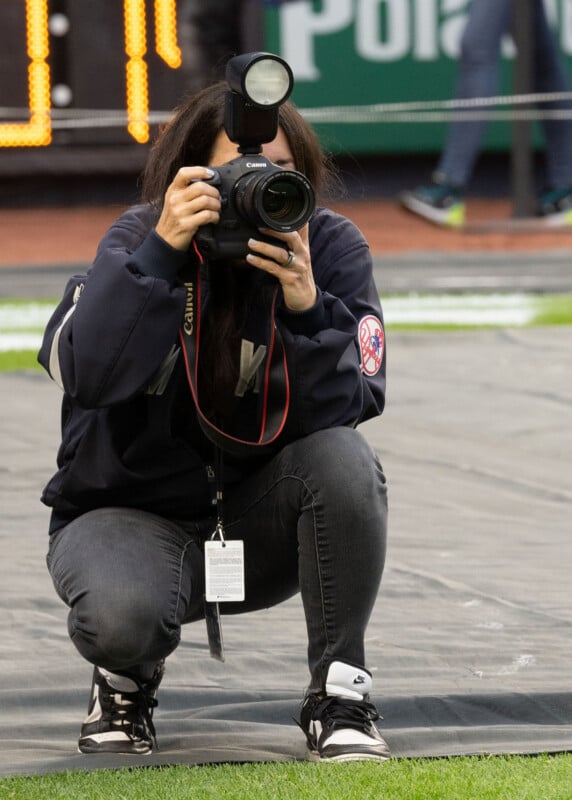 A person crouching on a sports field, wearing a dark jacket and sneakers, focuses a DSLR camera with a flash attached. They are capturing a moment, with blurred stadium elements in the background.