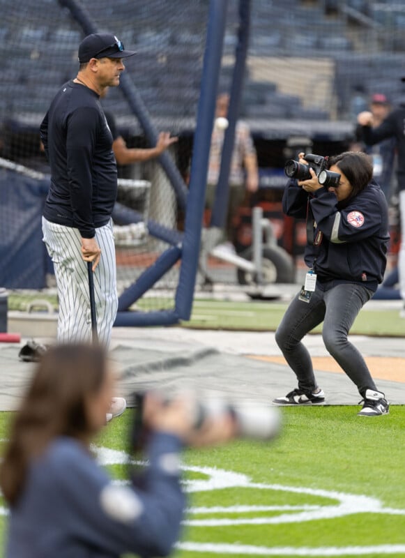 A person in a dark cap and baseball uniform is standing on a field, holding a bat. Another person is crouching and taking photos of them. The scene is set in a baseball stadium.