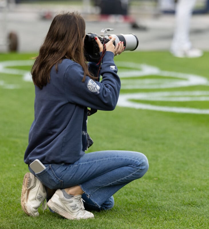 A person kneels on grass while taking a photo with a large camera. They are wearing a navy blue sweatshirt, jeans, and white sneakers. A phone is visible in their back pocket.