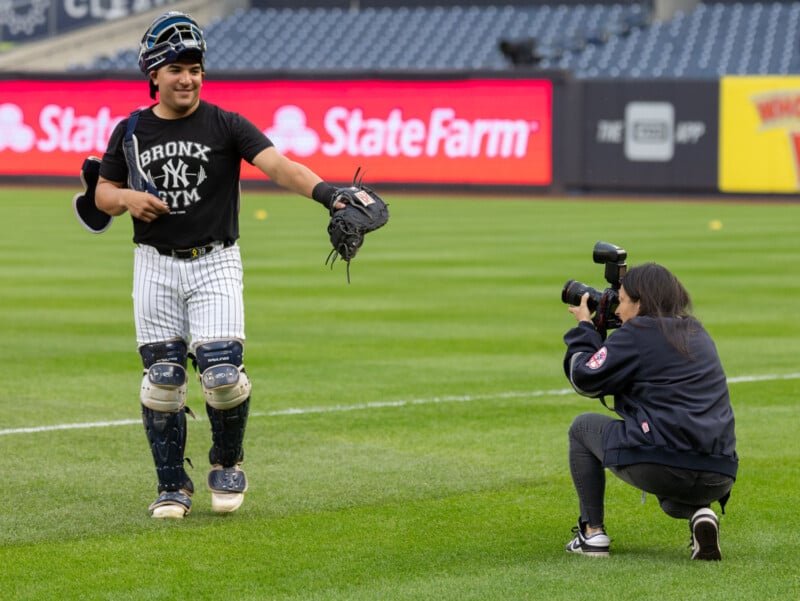 A baseball player in catching gear walks on a field, smiling and carrying a glove. A photographer kneels nearby, capturing the moment. The background features stadium advertisements.