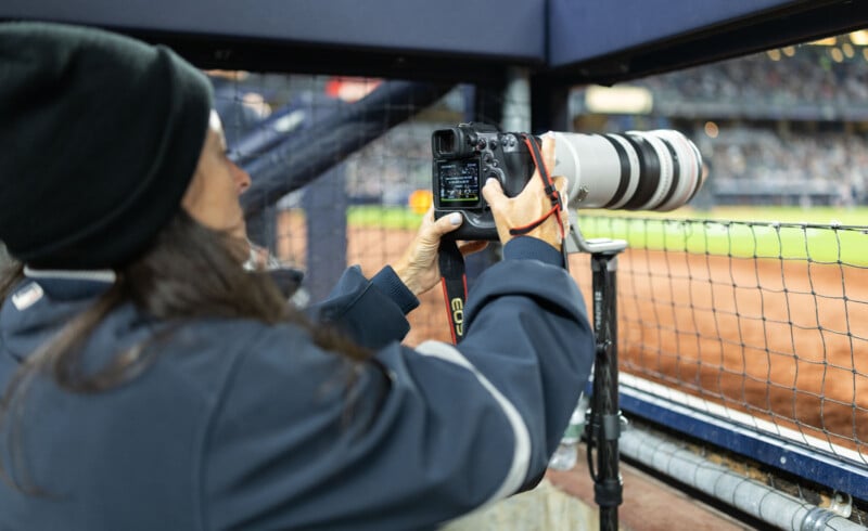 Person wearing a beanie and jacket uses a camera with a large zoom lens to photograph a baseball game from behind a wire fence. The camera is mounted on a tripod, and the baseball field is visible in the background.