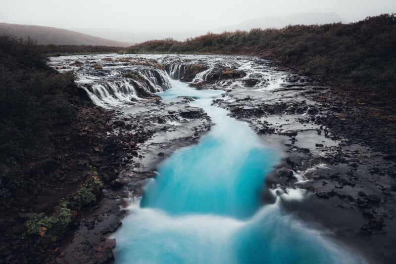 A breathtaking waterfall cascades over rocky ledges, with vivid turquoise water flowing through a rugged landscape surrounded by dense greenery and misty hills in the background.