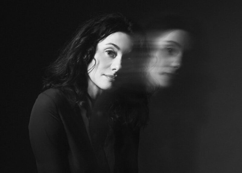 A monochrome depiction of a woman with long ebony hair, positioned against a dark backdrop. Her visage presents a ghostly double exposure effect, rendering a blurred replica to her right, suggesting a notion of motion or duality.