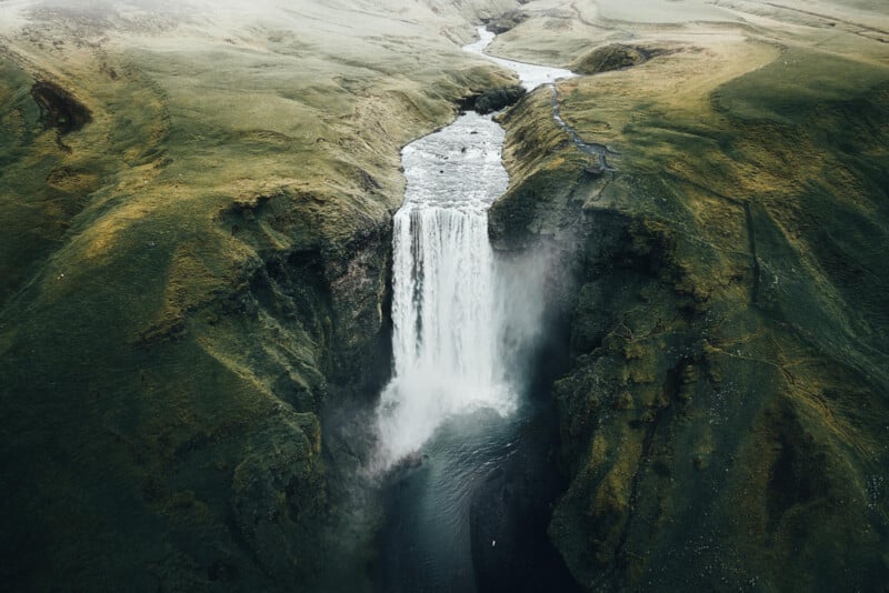 Top-down perspective of a majestic waterfall descending between verdant cliffs into a misty pool below, encircled by vibrant, rolling hills. A meandering river flows from the waterfall, cutting through the terrain.