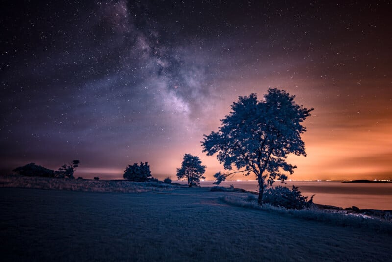 A tranquil nocturnal landscape showcasing a sky filled with stars, with the Milky Way observable. Trees are silhouetted near a peaceful water surface, and a warm orange hue on the horizon hints at a far-off city.