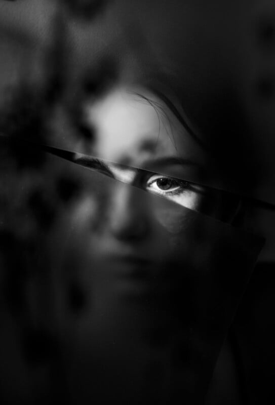 A close-up monochrome shot of a person's face shrouded in shadows and reflections. A beam of light highlights one eye, generating an enigmatic and dramatic effect. The arrangement features dark, indistinct textures throughout the image.