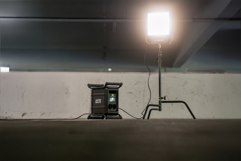 A portable light setup is positioned in a dimly lit parking garage. The light is attached to a tripod stand, and a control unit is placed nearby on a concrete surface. The environment appears industrial with visible wear on the walls.