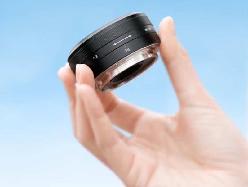 A hand is holding a camera lens against a clear blue sky. The lens is black with markings indicating a focal length of 1.8 and a focus range from 0.3 meters to infinity.