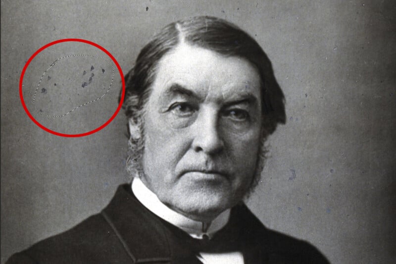 A black and white portrait of an older man with sideburns, wearing a dark coat and white shirt. A red circle highlights a section of the image with visible scratches or marks. The background is plain.