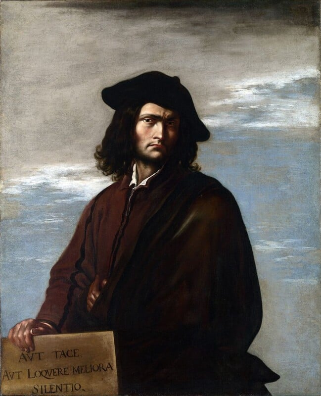 A man with lengthy hair and a dark hat stands against an overcast background, draped in a dark cloak. He holds a plaque inscribed with Latin text: "AUT TACE AUT LOQUERE MELIORA SILENTIO," meaning "Be silent, or let your words be better than silence.