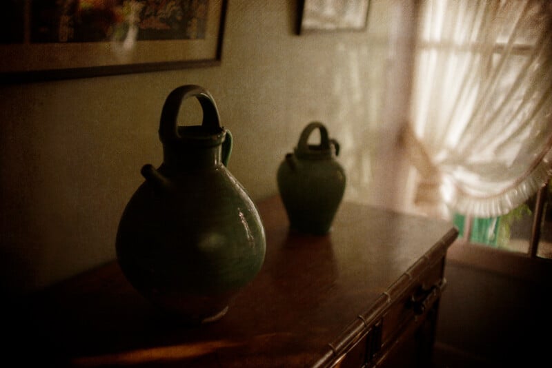 Two green pottery jugs are placed on a wooden table in a softly lit room. The background features a window with sheer curtains, creating a warm, cozy atmosphere. Framed pictures are partially visible on the wall.