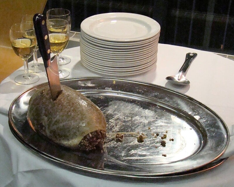 A haggis rests on a silver platter with a knife inserted into it. In the background, there are three glasses filled with white wine and a stack of white plates. A spoon lies beside the platter on a white tablecloth.