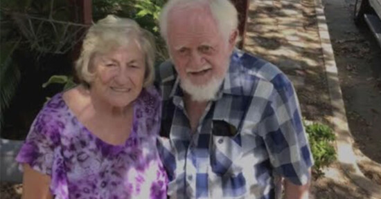 An elderly couple stands outdoors on a sunny day. The woman wears a purple patterned blouse, and the man wears a blue and white checkered shirt. They are smiling, surrounded by greenery and a sidewalk.