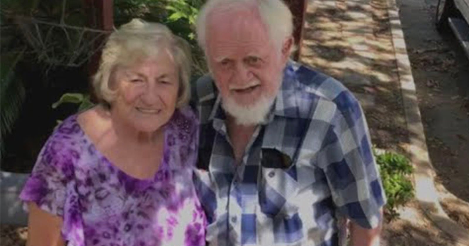 An elderly couple stands outdoors on a sunny day. The woman wears a purple patterned blouse, and the man wears a blue and white checkered shirt. They are smiling, surrounded by greenery and a sidewalk.