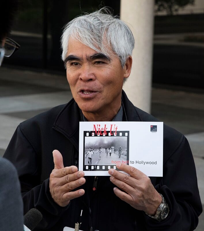 A man with gray hair is holding a book titled "Nick Ut: From Hell to Hollywood." The cover displays a black and white photo strip. He appears to be speaking and gesturing outdoors.