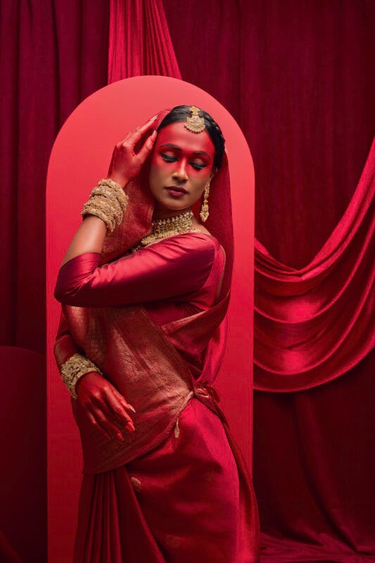 A person poses gracefully against a rich, burgundy backdrop, adorned in an elegant traditional outfit with gold jewelry, including a headpiece and bangles. The setting features flowing fabric and an arch, enhancing the luxurious ambiance.