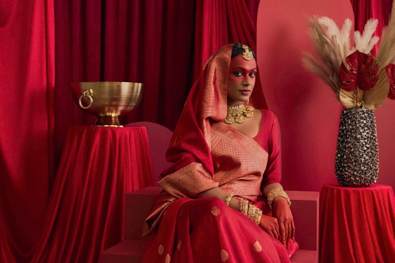 A woman in a traditional attire, featuring an ornate gold headpiece and jewelry, sits against a backdrop of rich red drapes. She is surrounded by elegant decor including a brass bowl and a vase with feather arrangements.