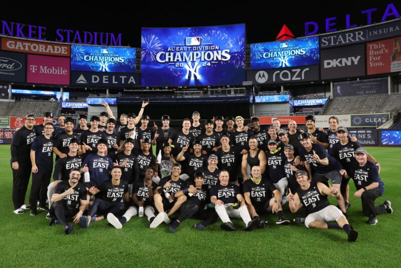 A baseball team poses for a celebratory group photo on a field. They are wearing matching shirts with "EAST" on them. A large stadium screen shows "AL East Division Champions 2024" with fireworks graphics in the background.