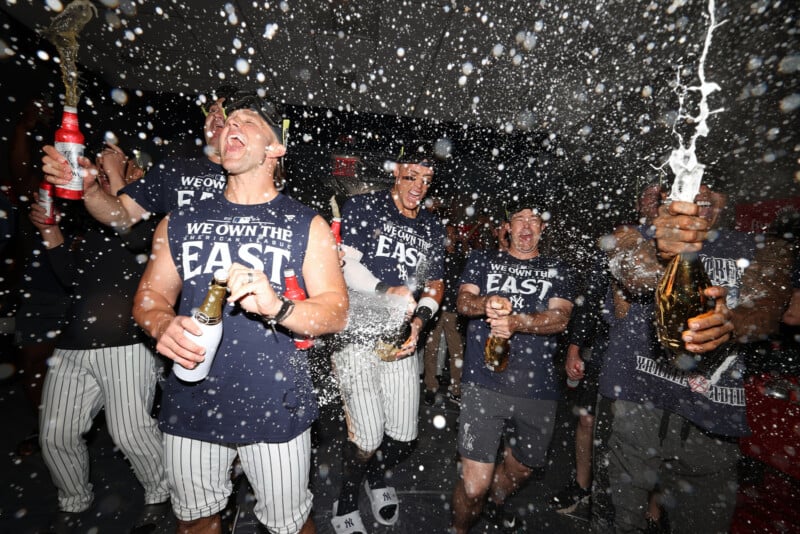 Baseball players in a locker room celebrate with champagne, wearing shirts that say "We Own The East." They are smiling, laughing, and spraying each other with bottles, with champagne droplets captured in mid-air.