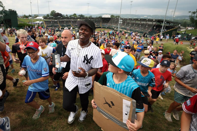 A man in a Yankees jersey happily walks through a grassy area surrounded by a crowd of smiling children and adults, some wearing baseball apparel. A stadium and playing fields are visible in the background.