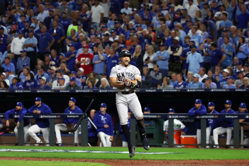 A baseball player in a New York uniform jumps in celebration on a field, surrounded by enthusiastic fans in the stands. The opposing team, in blue jerseys, sits in the dugout watching the action.