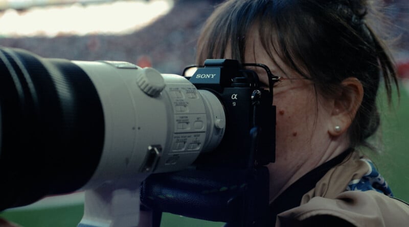 A person using a Sony camera with a large telephoto lens looks through the viewfinder. The background is blurred, suggesting they are at an outdoor event. The camera and lens are prominent in the foreground.