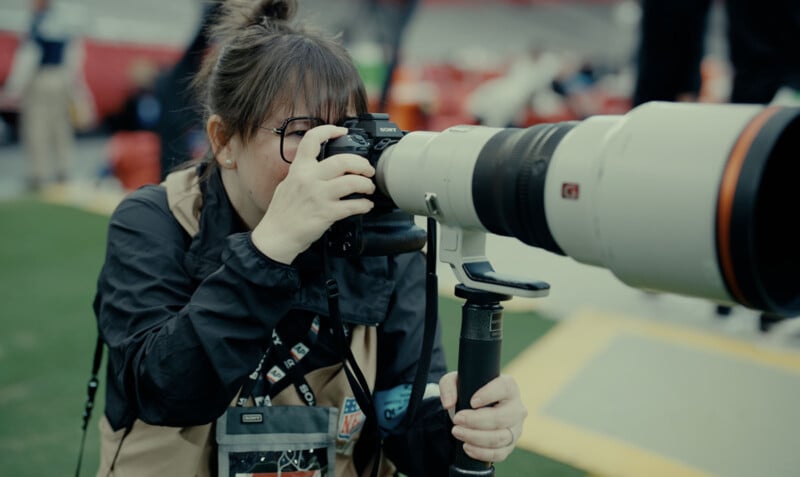 A photographer wearing a brown vest is intently taking a photo with a professional camera and large telephoto lens at an outdoor event. She appears focused, and there are blurred figures in the background.