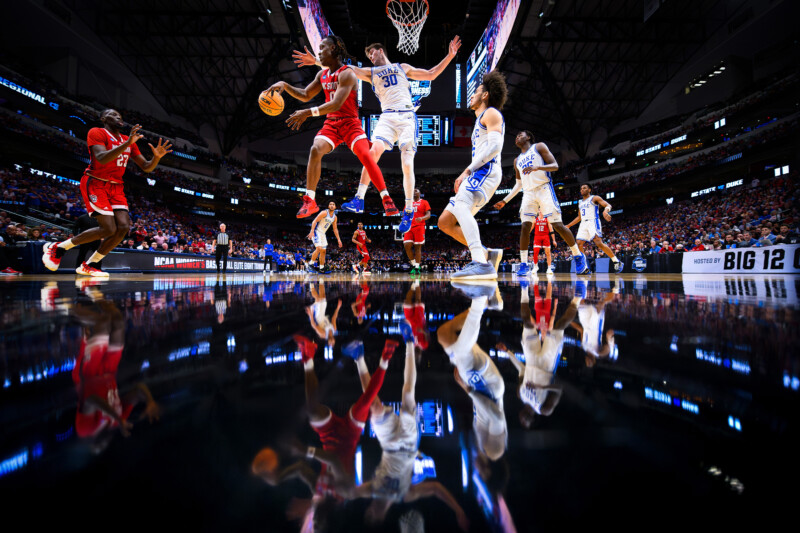 Basketball players in red and blue jerseys are caught in action on the court amidst a packed stadium. The glossy floor reflects the captivating scene, encapsulating the intensity of the moment.