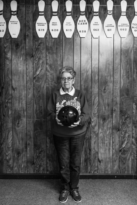 A black and white image of an older person standing in front of a wooden wall, holding a bowling ball. The wall displays several bowling pins with names and numbers written on them. The person is wearing glasses, a sweater, jeans, and sneakers.