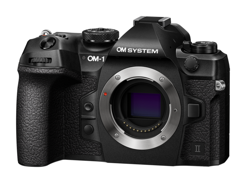 The image shows an OM System camera body. It's black with textured grip and various control buttons. The lens mount is exposed, revealing the sensor. The brand name is visible on the top left of the camera.