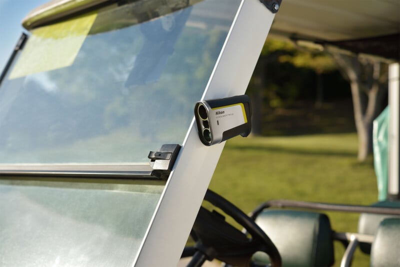 A Nikon golf rangefinder is mounted on the windshield frame of a golf cart. The cart is on a golf course, with green grass and trees visible in the background under a clear sky.