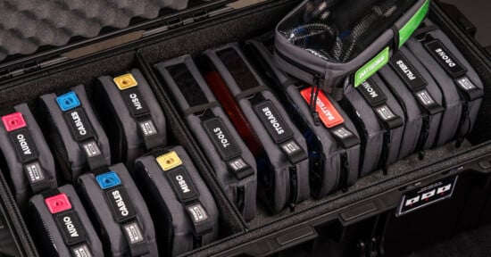 A neatly organized case holds various labeled electronic gear pouches. Labels include audio, cables, tools, storage, batteries, filters, and more. The pouches are color-coded and fit precisely in the protective foam interior.