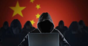 A person in a hooded sweatshirt sits in front of a laptop with a dimly lit, shadowy face. Behind them, the Chinese flag is prominently displayed. The background shows several similar figures in a row.