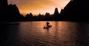 A person stands on a small boat with a cormorant bird at sunset. They are silhouetted against an orange sky and surrounded by the jagged peaks of karst mountains, creating a tranquil scene on the water.