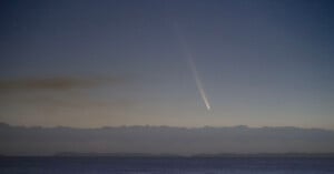 A comet with a long, bright tail streaks across a starry night sky above the horizon, casting a serene glow over the calm ocean below. Dark silhouettes of distant landmasses are visible at the horizon line.